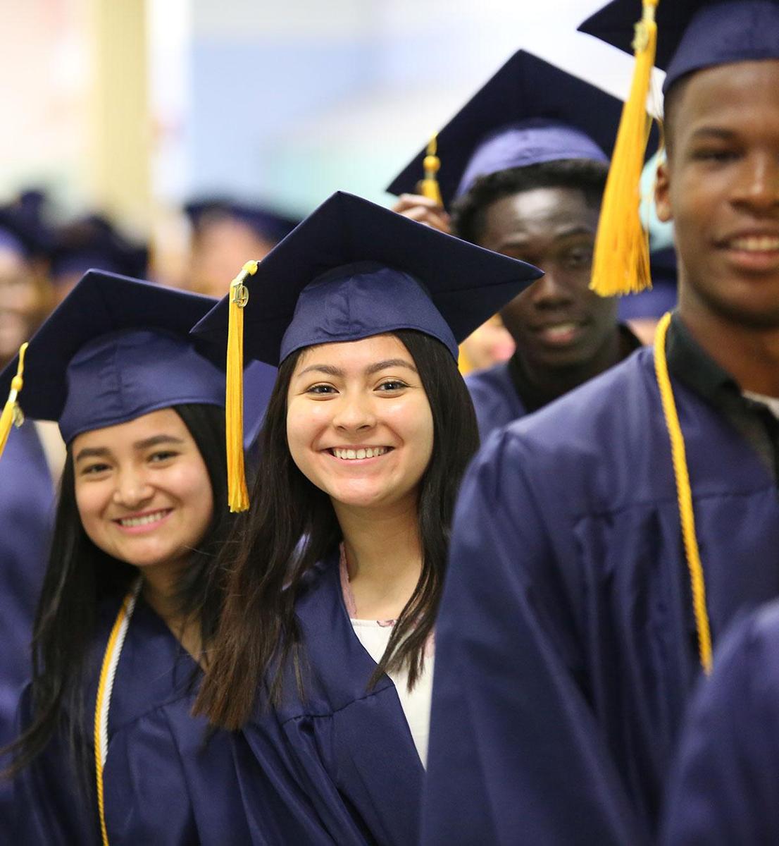 Smiling high school graduates in navy blue caps and gowns standing in line during a graduation ceremony.